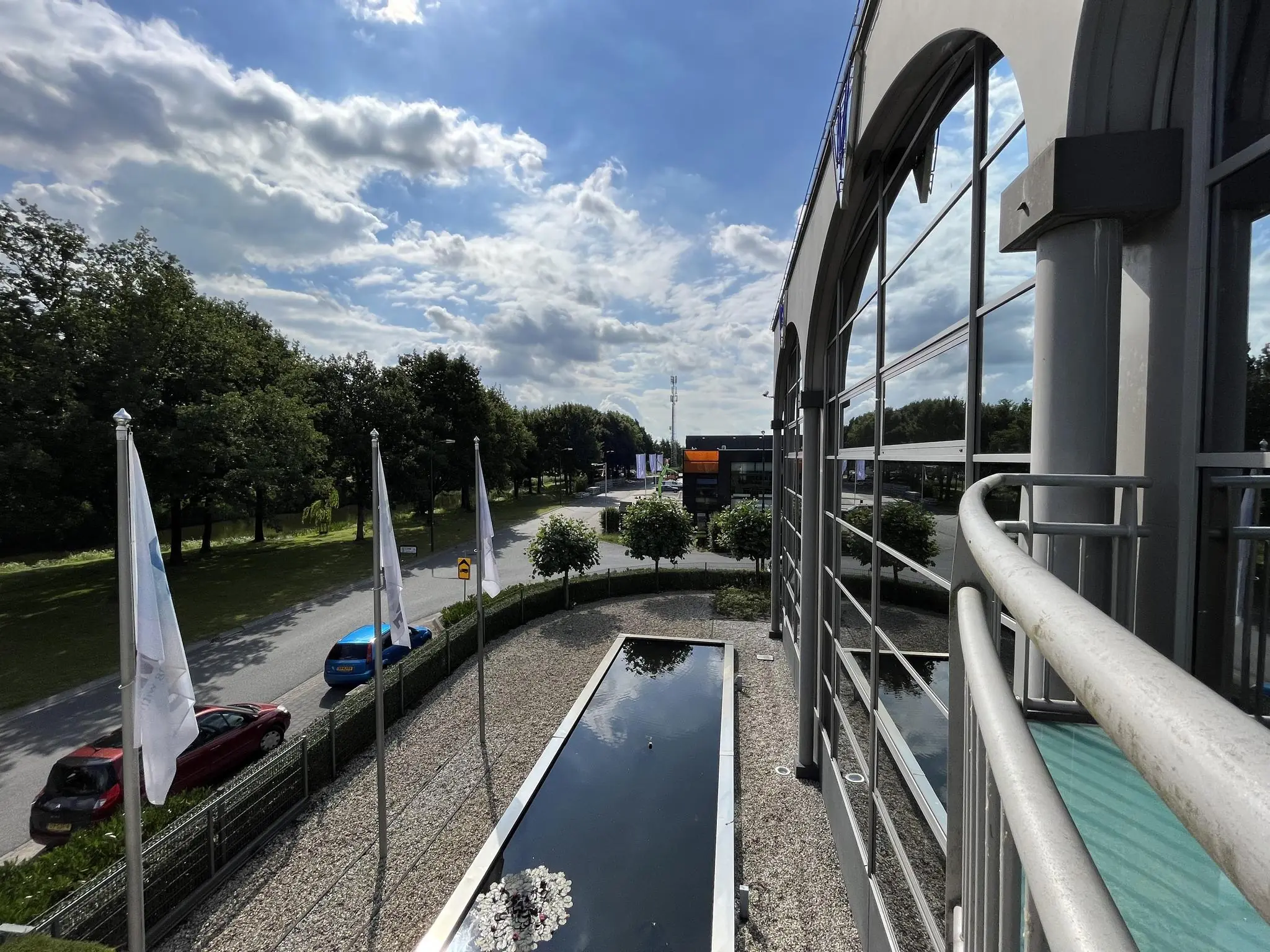Zijaanzicht van een modern kantoorgebouw aan de Oostergracht met vlaggen, vijver en bomen langs de weg onder een deels bewolkte hemel.