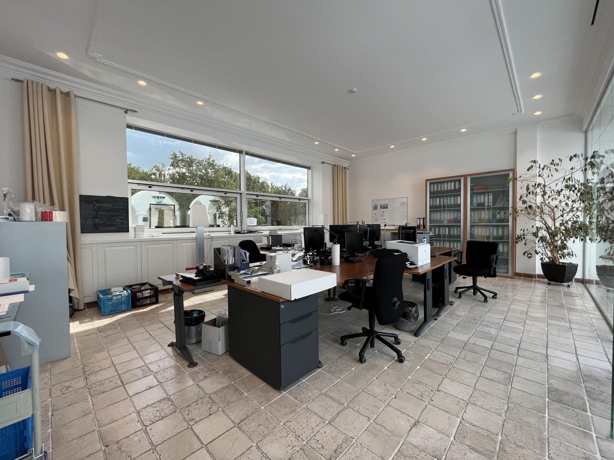 Een licht en ruim kantoor met bureaus, stoelen en grote ramen met uitzicht op groen.