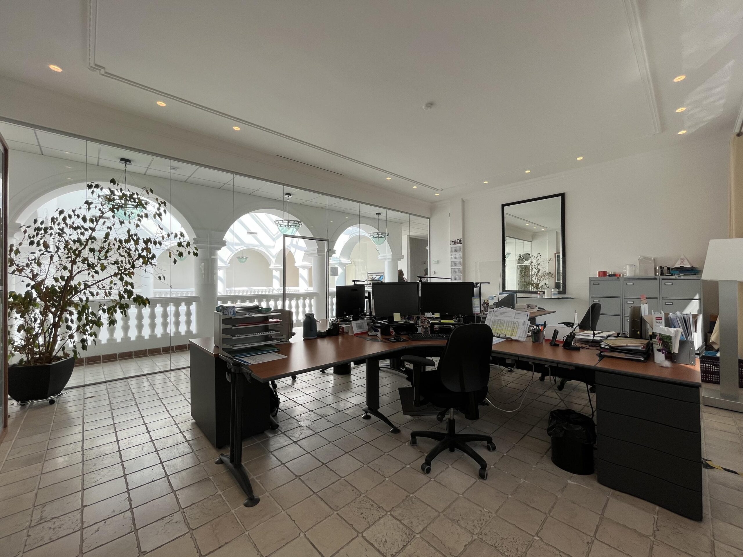 Een modern kantoor met bureaus, stoelen, en computers, met glazen wanden die uitkijken op een marmeren balkon met bogen.