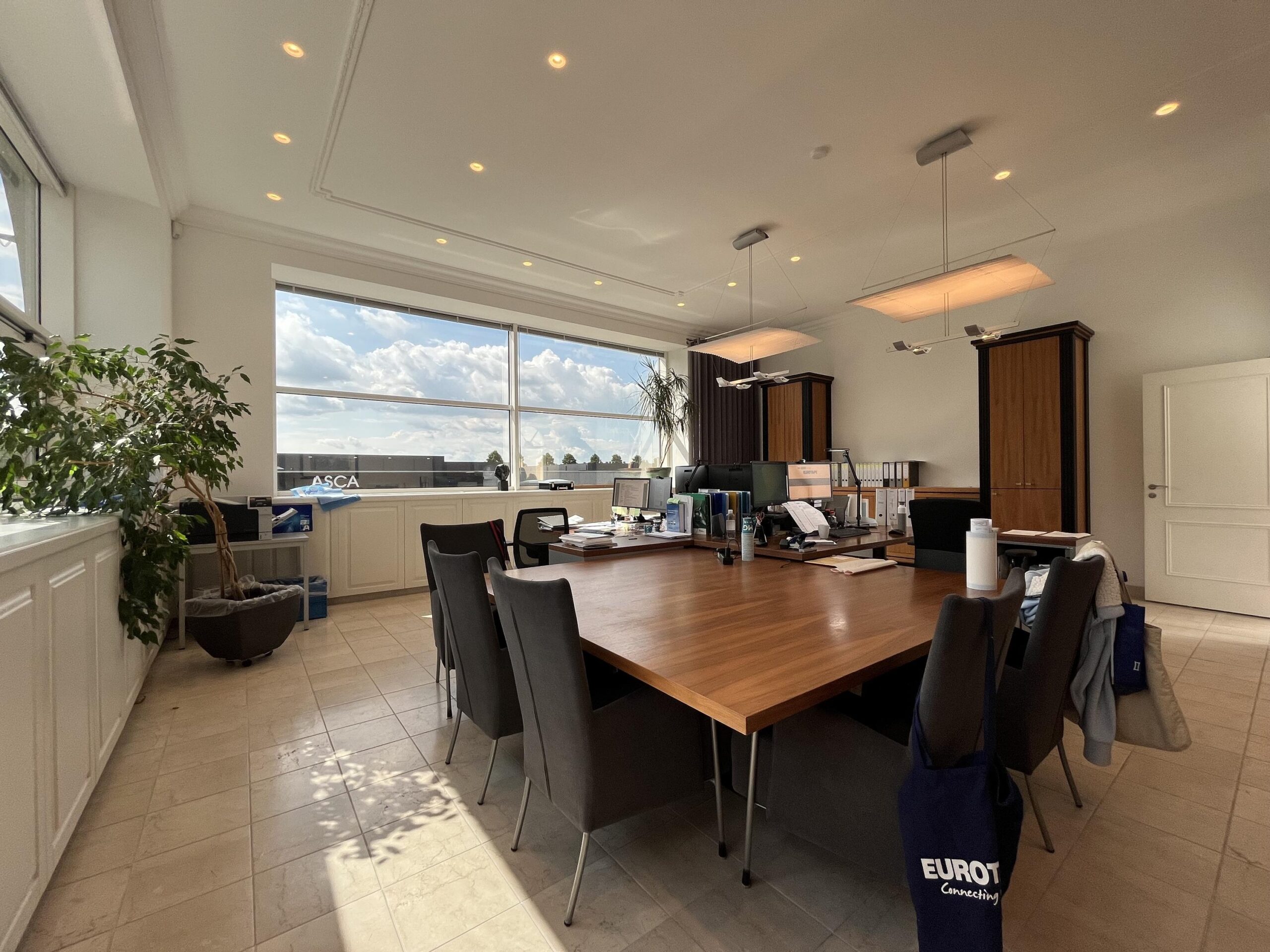 Een moderne vergaderruimte met een grote houten tafel en stoelen, verlicht door natuurlijk licht vanuit een groot raam.