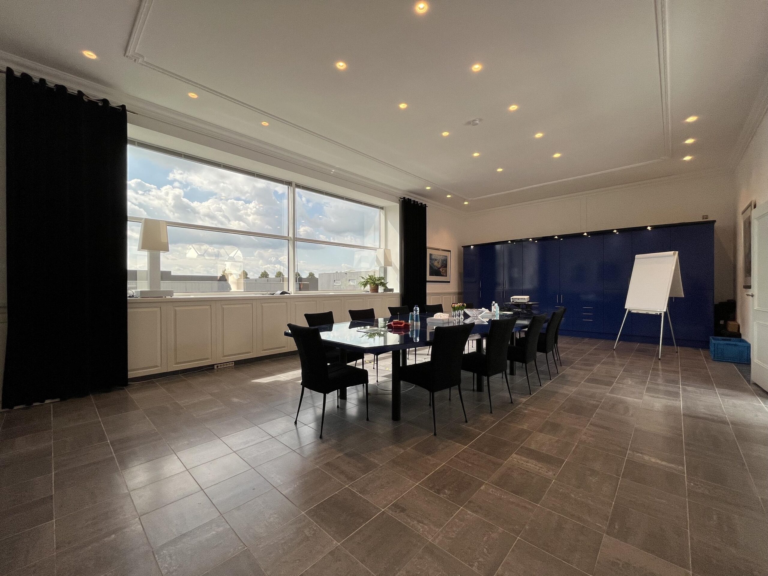 Vergaderruimte met een grote tafel, stoelen, flip-over, en een raam met uitzicht op een heldere lucht.