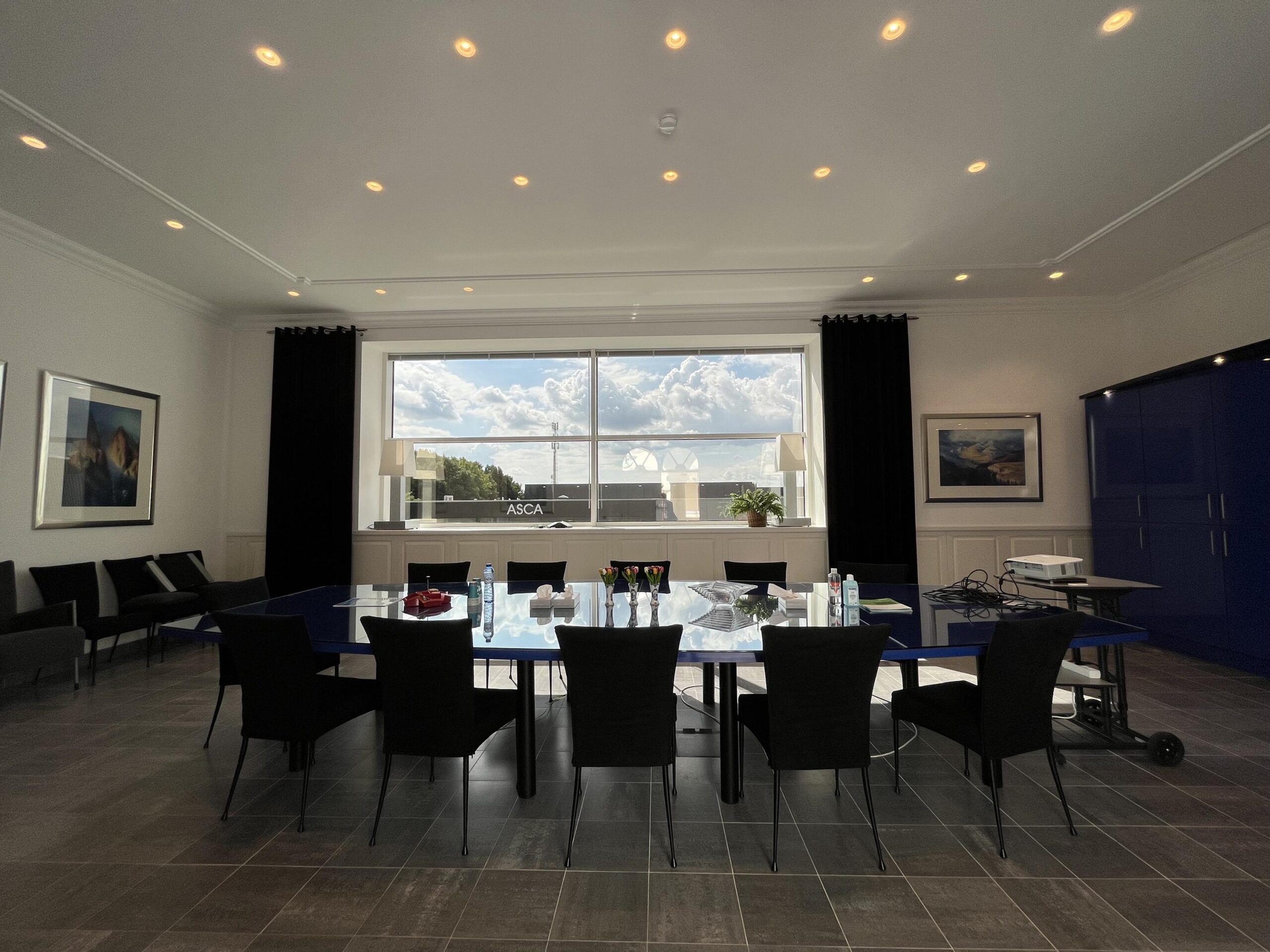 Een vergaderruimte met een grote tafel, stoelen, en een raam met uitzicht op de lucht en wolken.