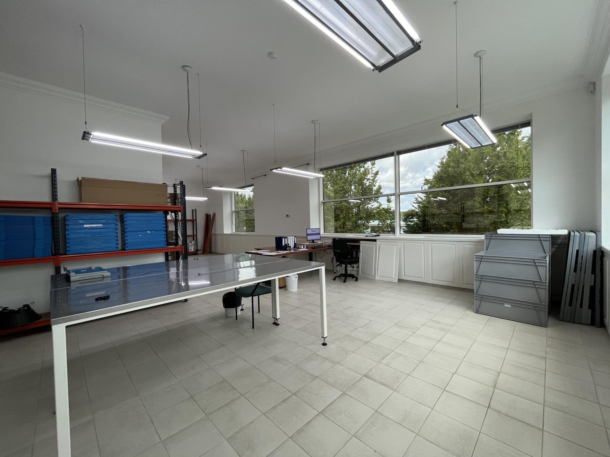 Een moderne, lichte werkruimte met tafels, opslagrekken en grote ramen met uitzicht op bomen.