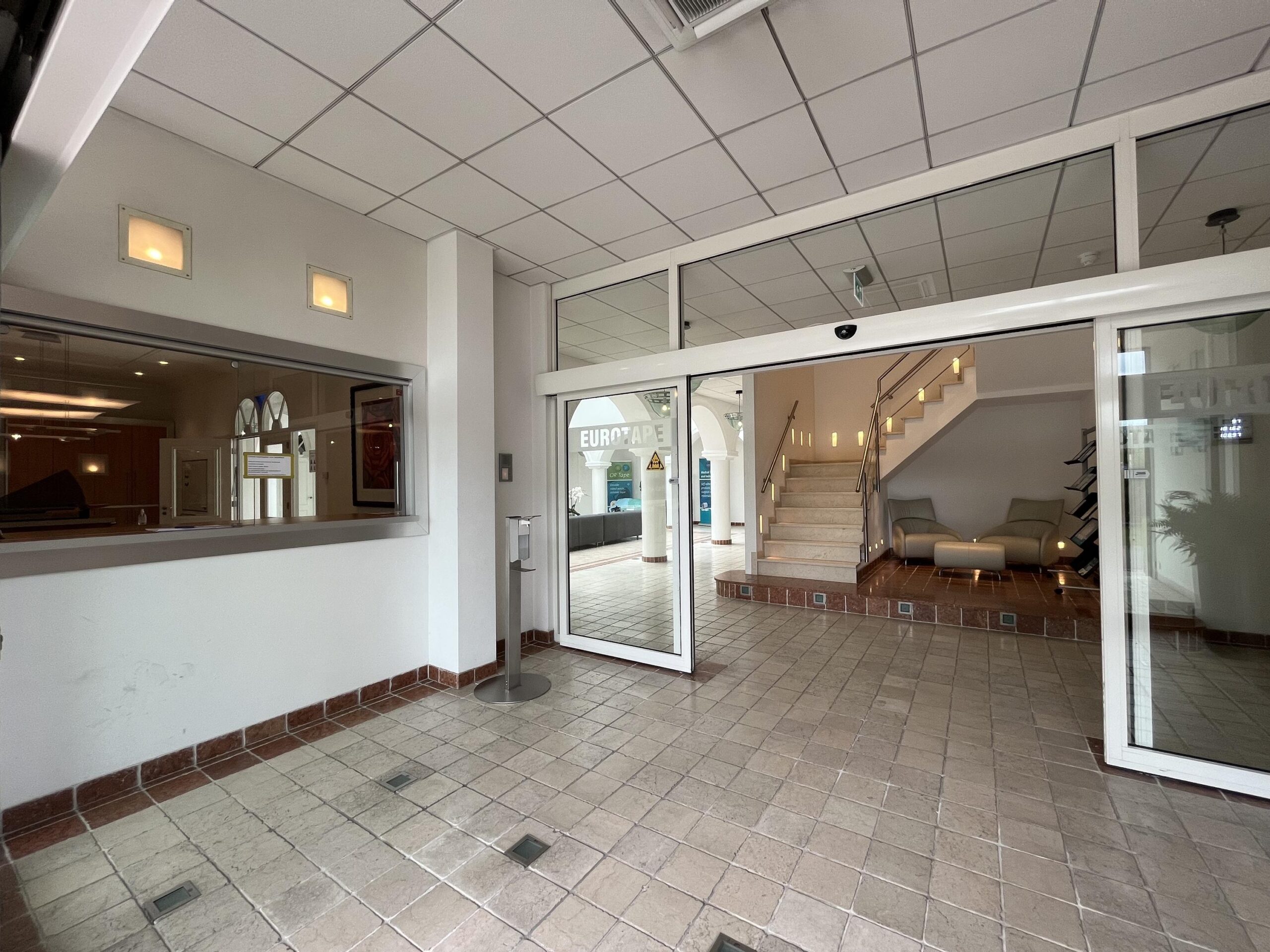 Ingang van een modern kantoorgebouw met glazen deuren, een receptie, en een trap met een zitgedeelte.