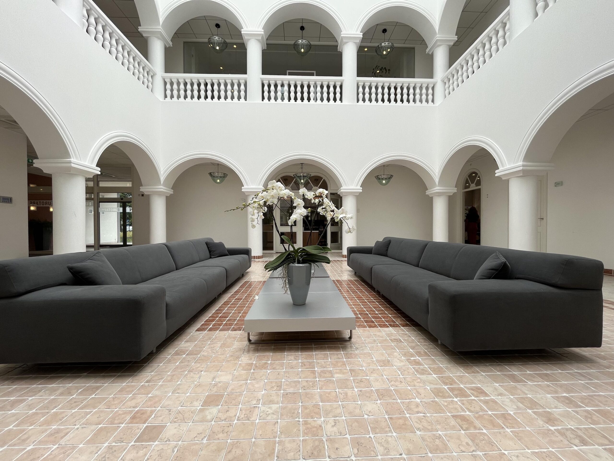 Een lichte, open ruimte met hoge witte bogen, grijze banken en een tafel met een plantenbak.