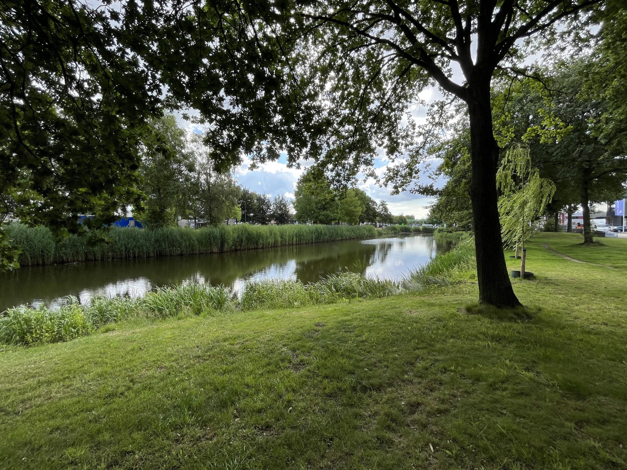 Een rustige gracht omgeven door groene bomen en gras, met weerspiegeling van de lucht in het water.