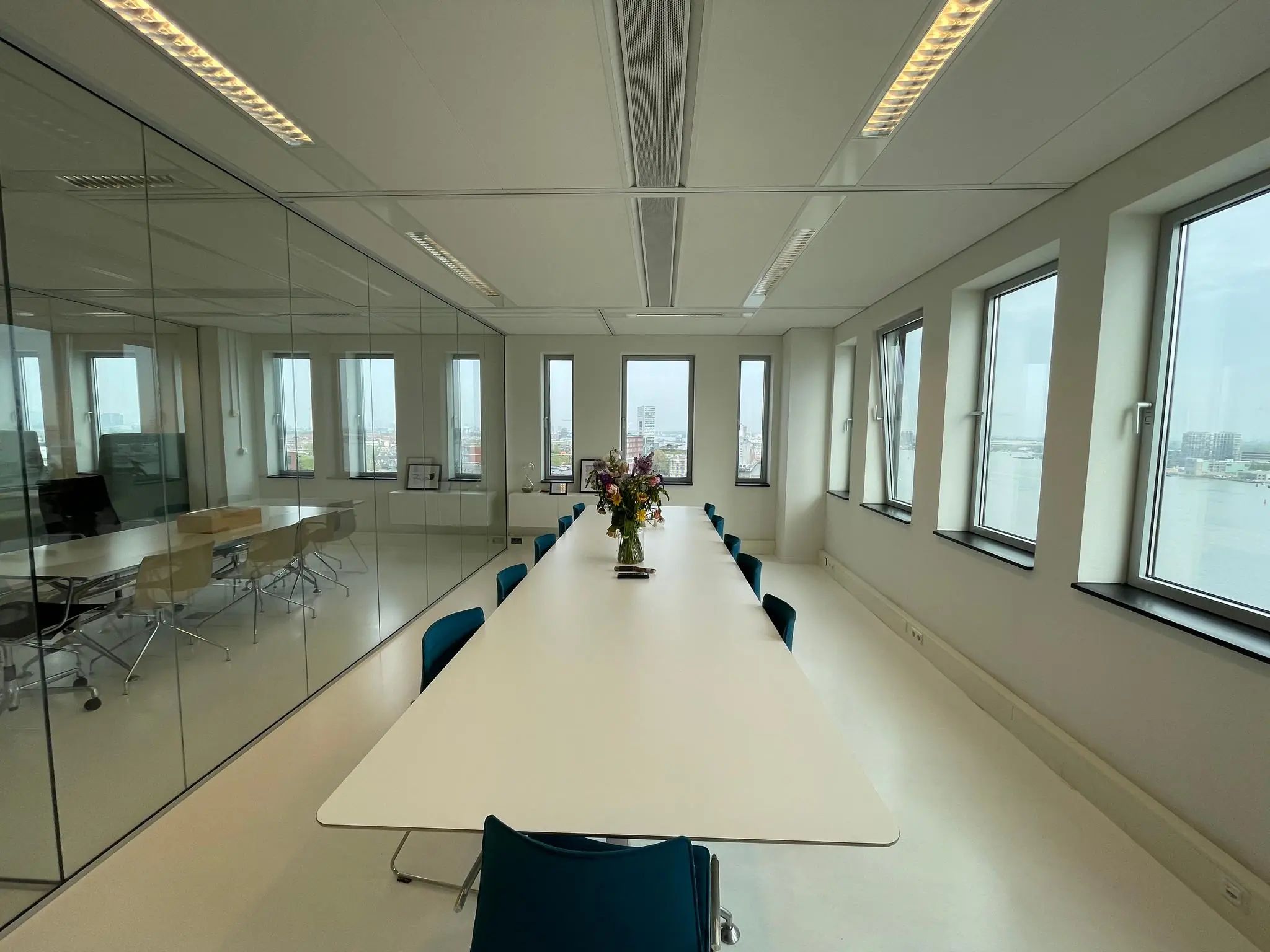 Vergaderruimte met lange tafel, blauwe stoelen, bloemen in het midden en uitzicht op het IJ in Amsterdam vanaf het IJdok.