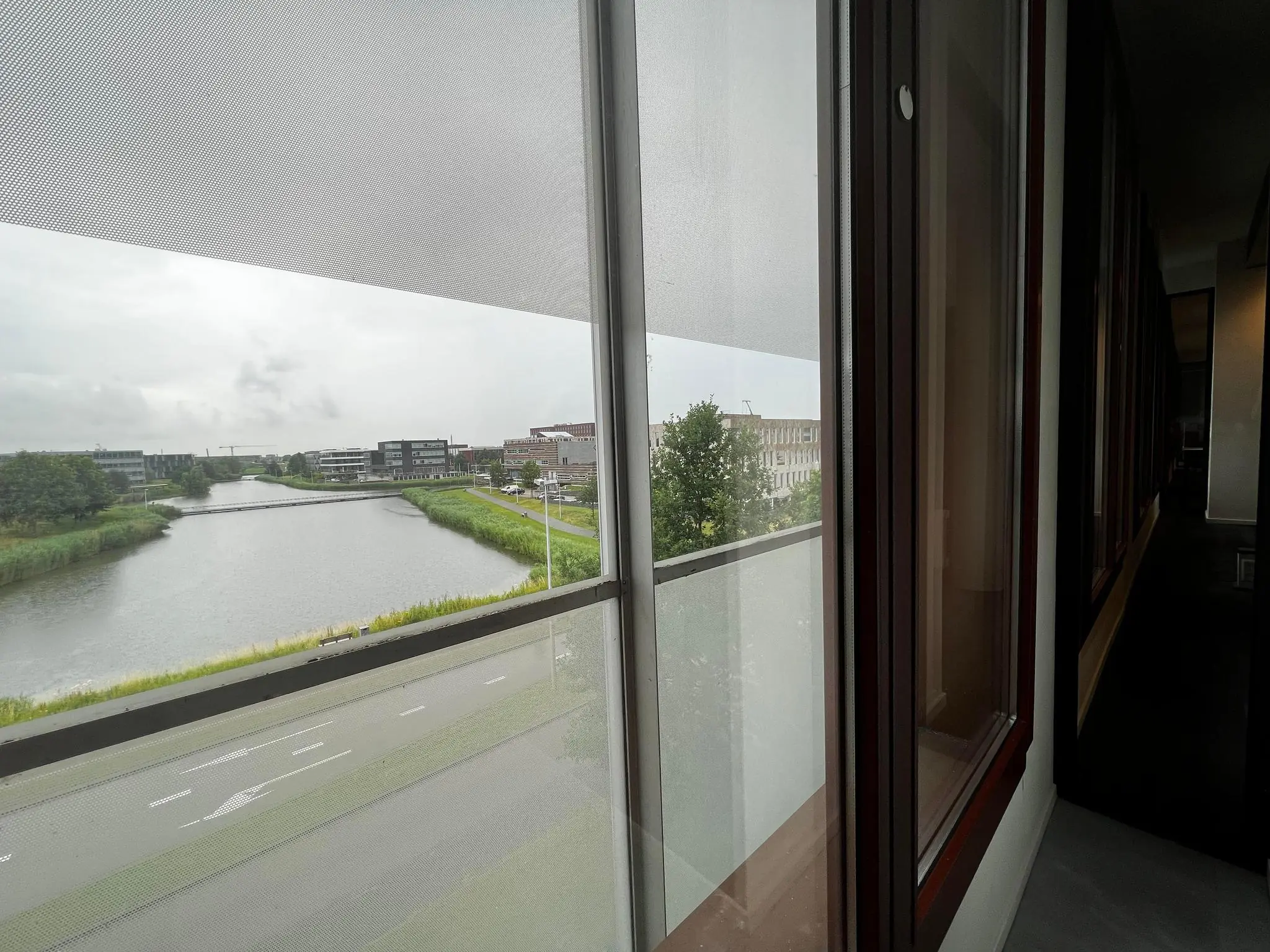 Uitzicht vanaf een gang met ramen op de Hondiuslaan, met zicht op een waterloop, brug en omliggende kantoorgebouwen.