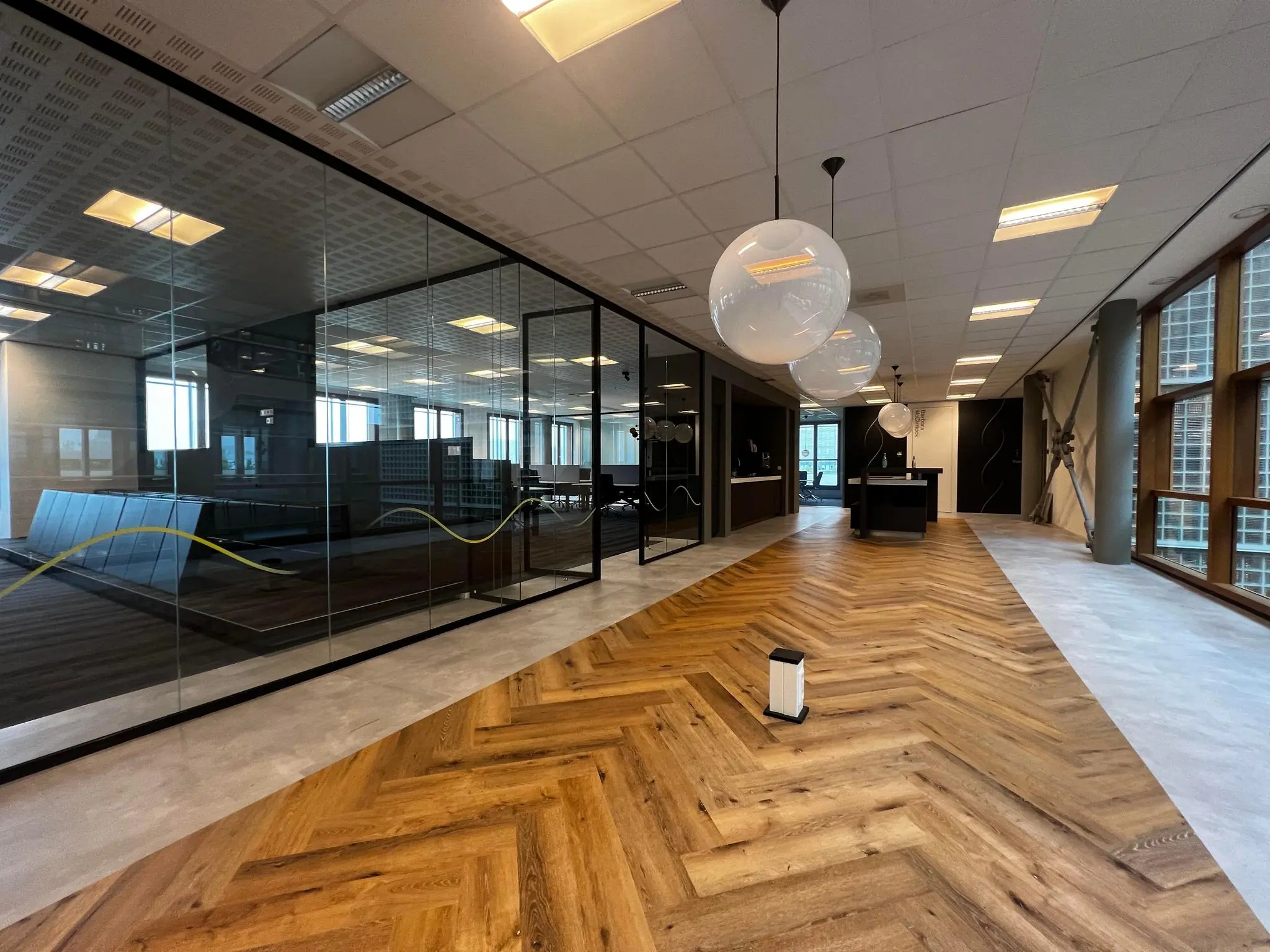Moderne kantoorgang aan de Hondiuslaan met visgraat houten vloer, glazen wanden en hangende ronde lampen.