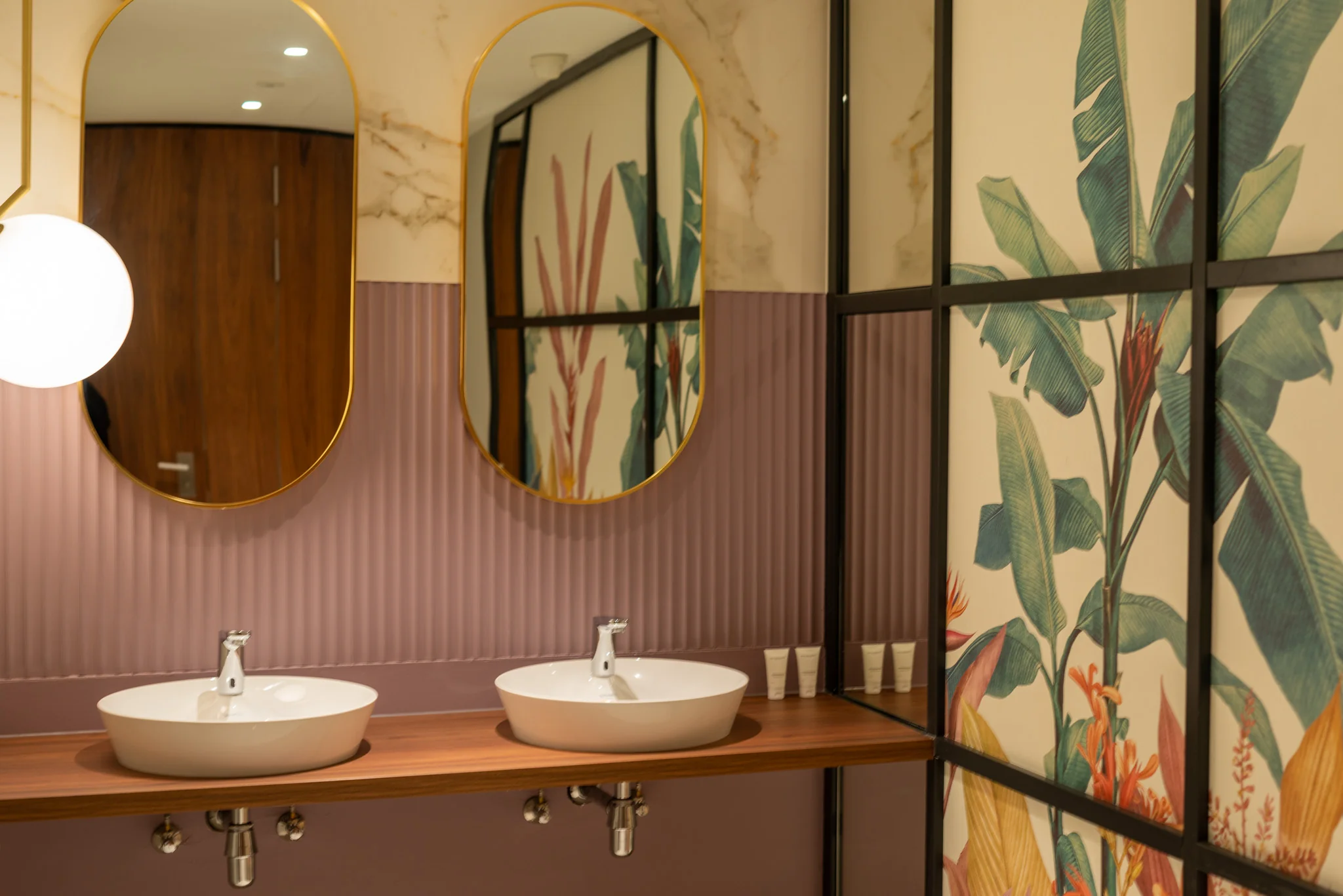 Moderne badkamer met twee ovalen spiegels, wasbakken en een kleurrijke wand met botanisch design.