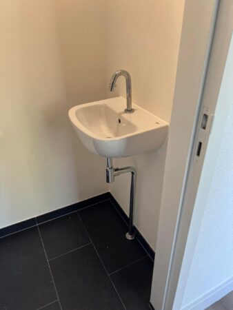 Wit hoekfonteintje met chromen kraan en afvoer in een strak afgewerkte ruimte met zwarte vloertegels.