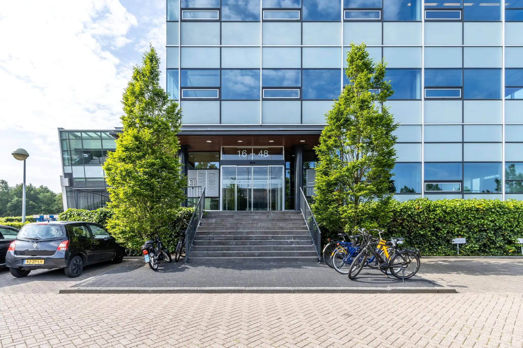 Ingang van een modern kantoorgebouw aan de Rijnzathe 16-48 met trap, fietsen en geparkeerde auto’s.