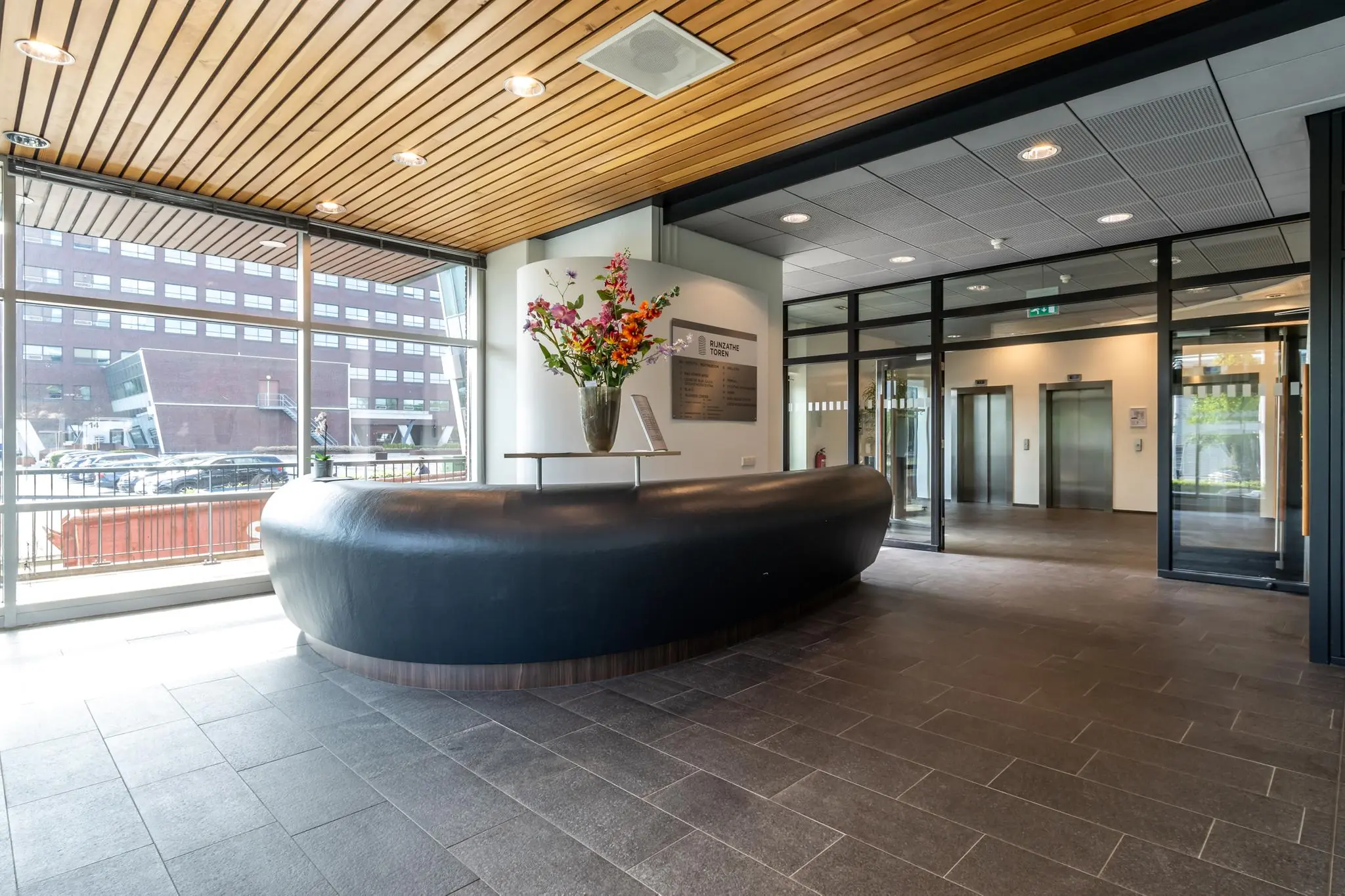 Moderne ontvangstbalie met bloemen in de hal van kantoorgebouw Rijnzathe, met zicht op liften en grote raampartijen.