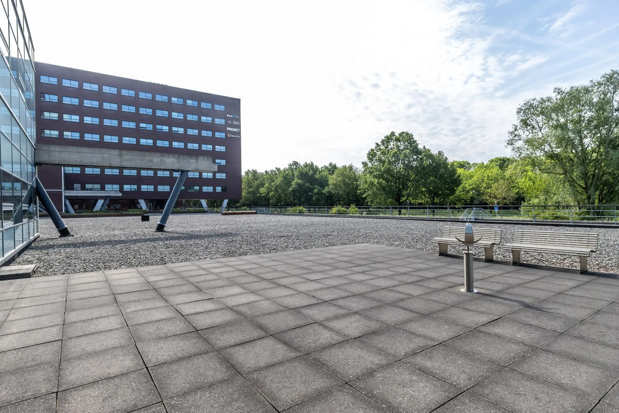 Modern kantoorgebouw aan de Rijnzathe met betegeld plein, zitbanken en uitzicht op een groene boomrand.