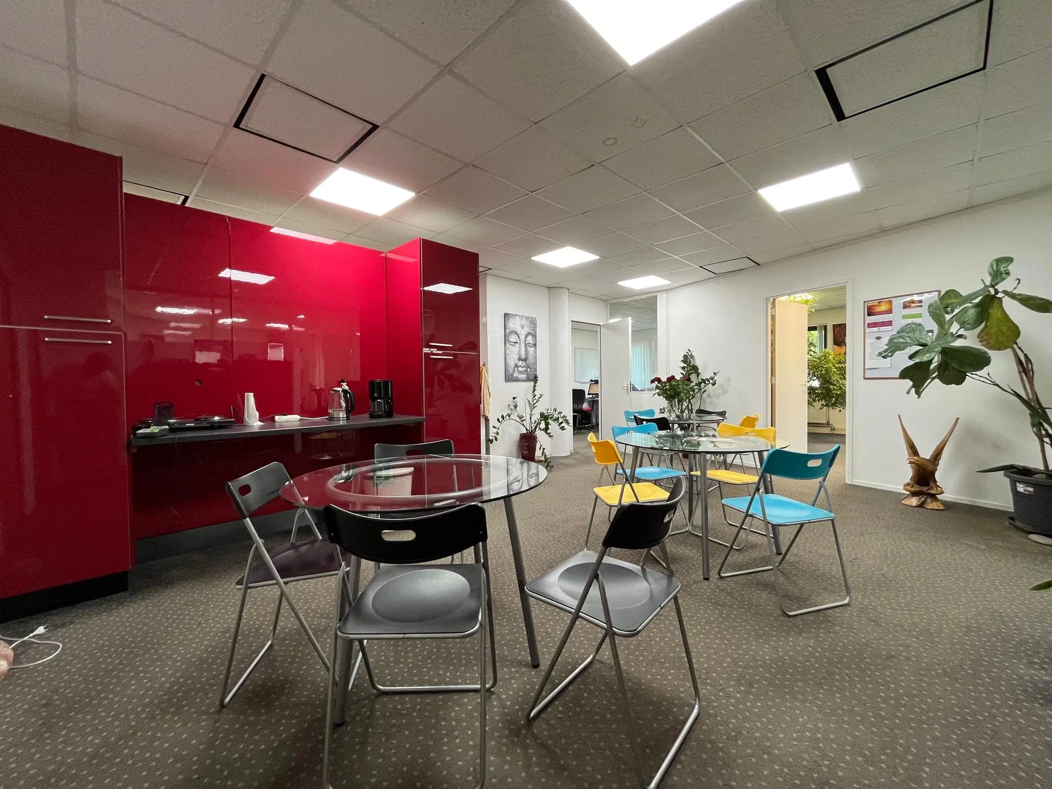 Kantineruimte met glazen tafels, gekleurde stoelen en een felrode keukenwand in een kantoor aan de Koningsweg.
