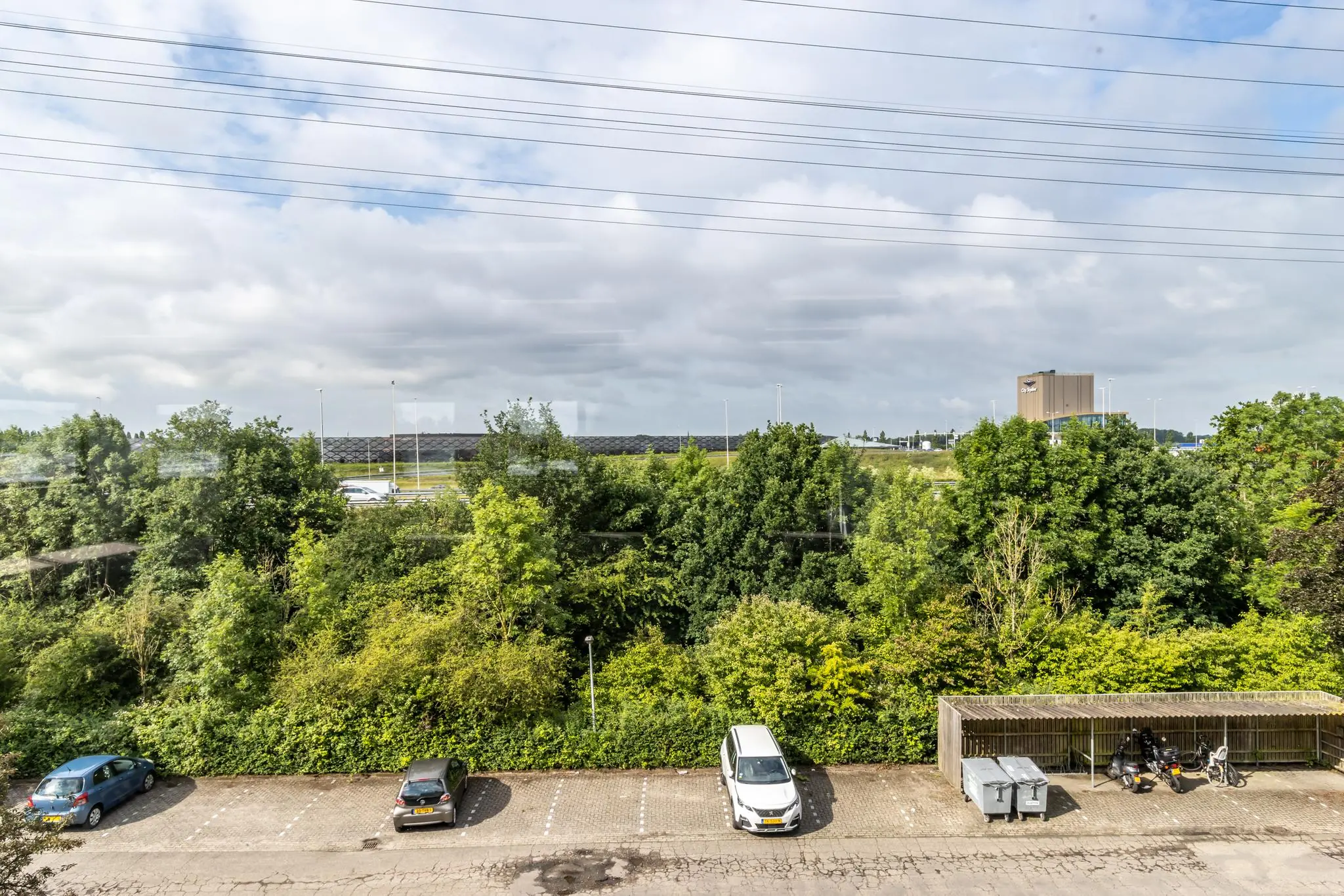 Parkeerplaats met enkele auto's en een fietsenstalling aan de Zonnebaan, met op de achtergrond bomen, een snelweg en een bedrijfsgebouw.