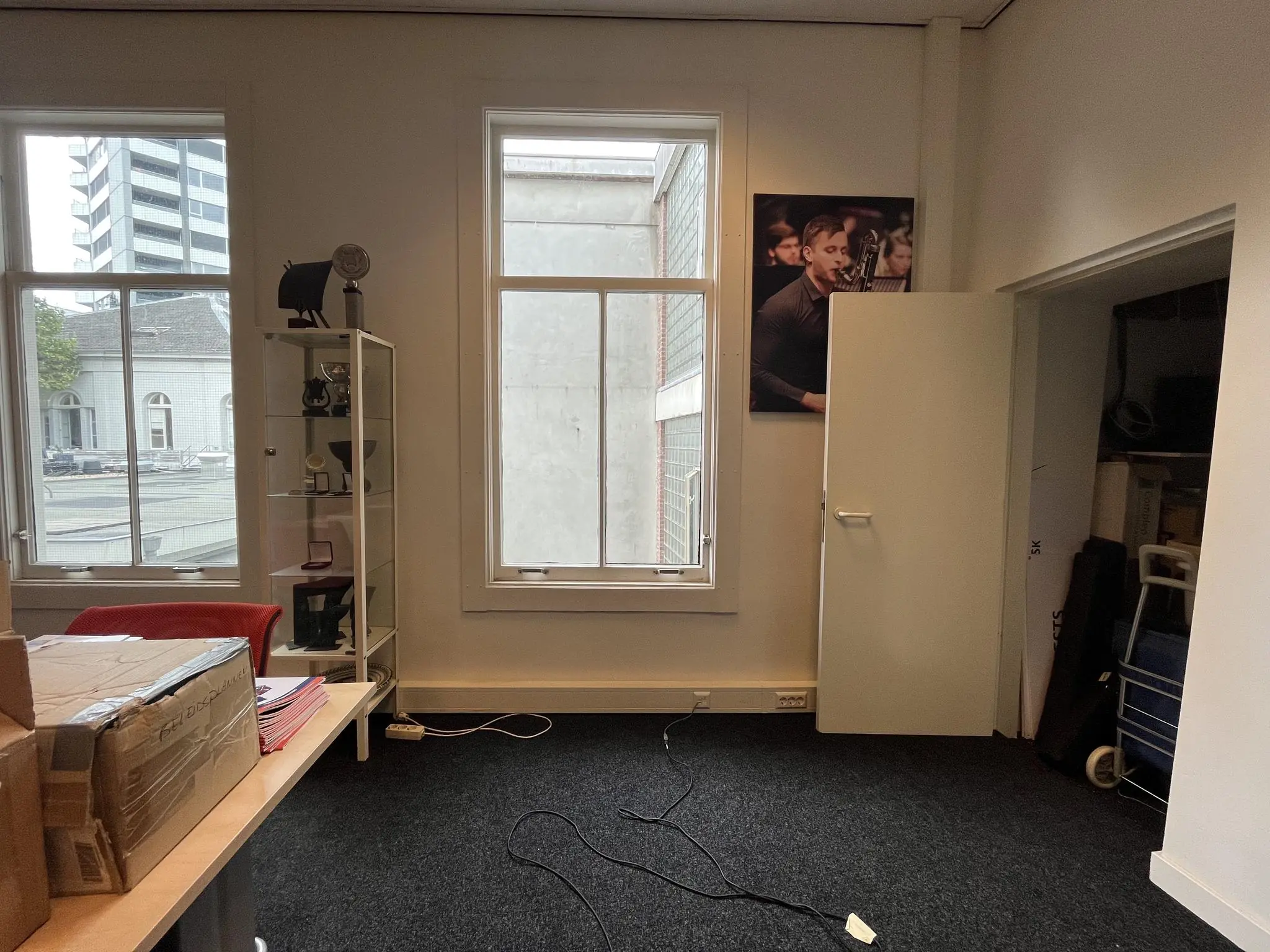 Kantoorruimte met twee ramen, een vitrinekast, dozen op een bureau en een open deur naar een opslagruimte, genomen aan de Ganzenmarkt.