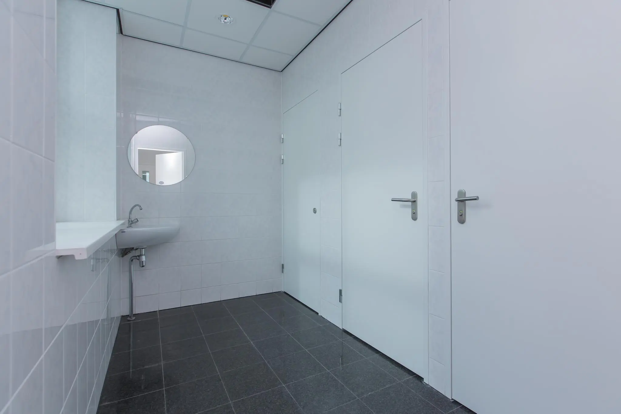 Witte, betegelde toiletruimte in kantoorgebouw Rhijnspoor met twee deuren, een wastafel en een ronde spiegel.