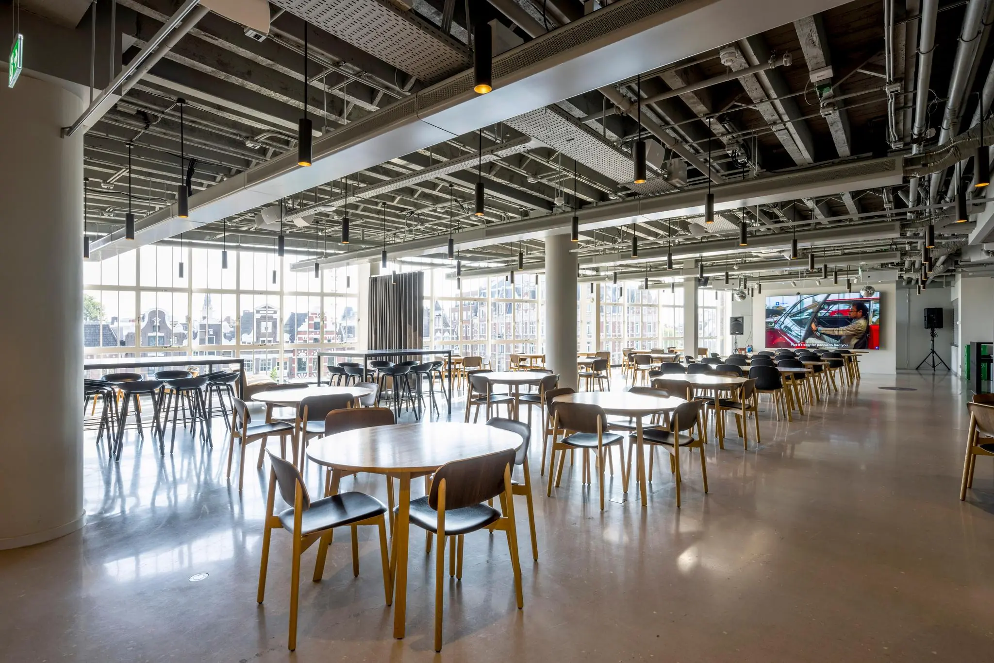 Moderne, ruime bedrijfskantine aan het Rokin met grote ramen, houten tafels en stoelen, en een scherm aan de achterwand.