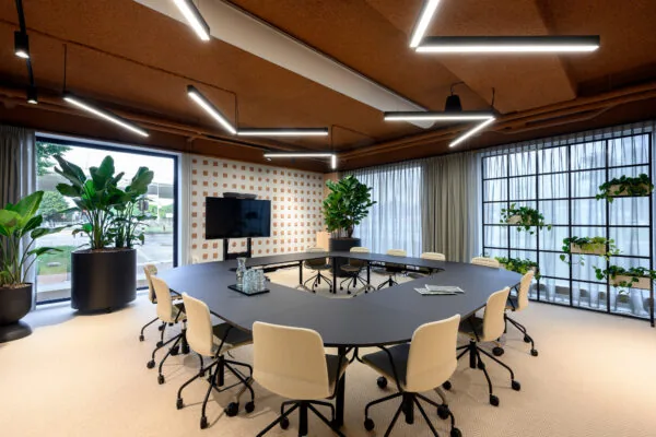 Moderne vergaderruimte met U-vormige tafelopstelling, beige stoelen, grote planten en natuurlijk licht.