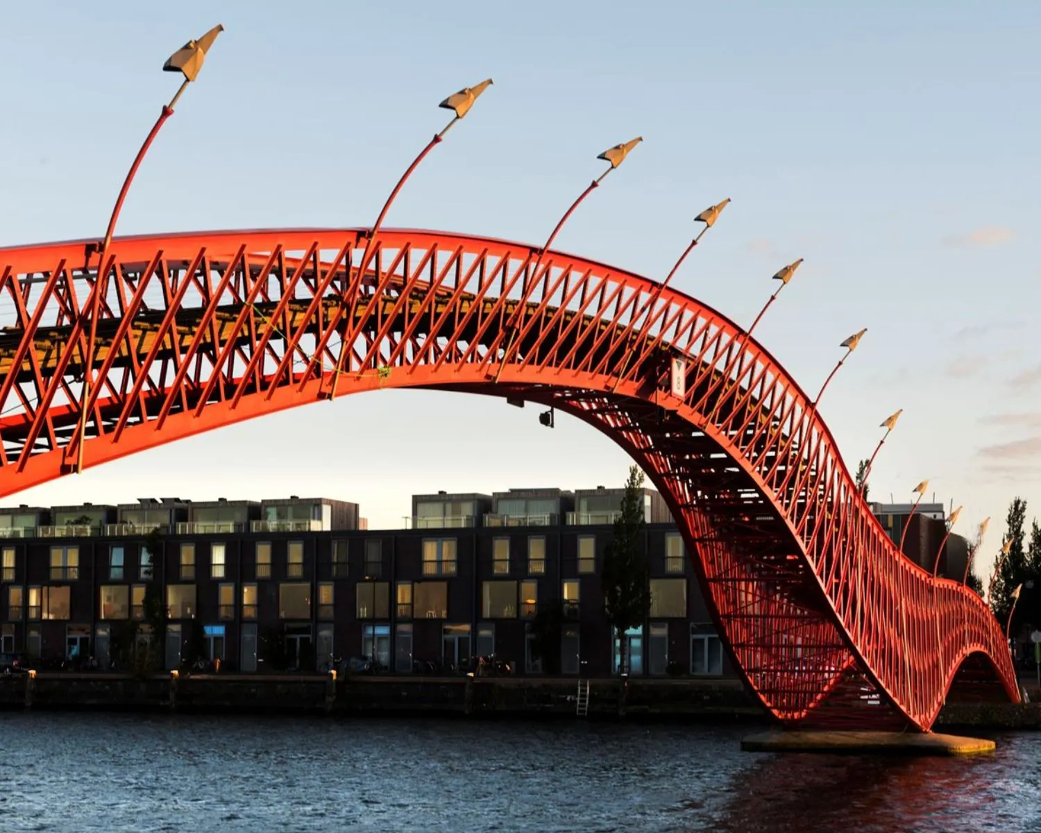 Golvend rode Pythonbrug in Amsterdam steekt over het water met moderne gebouwen op de achtergrond.
