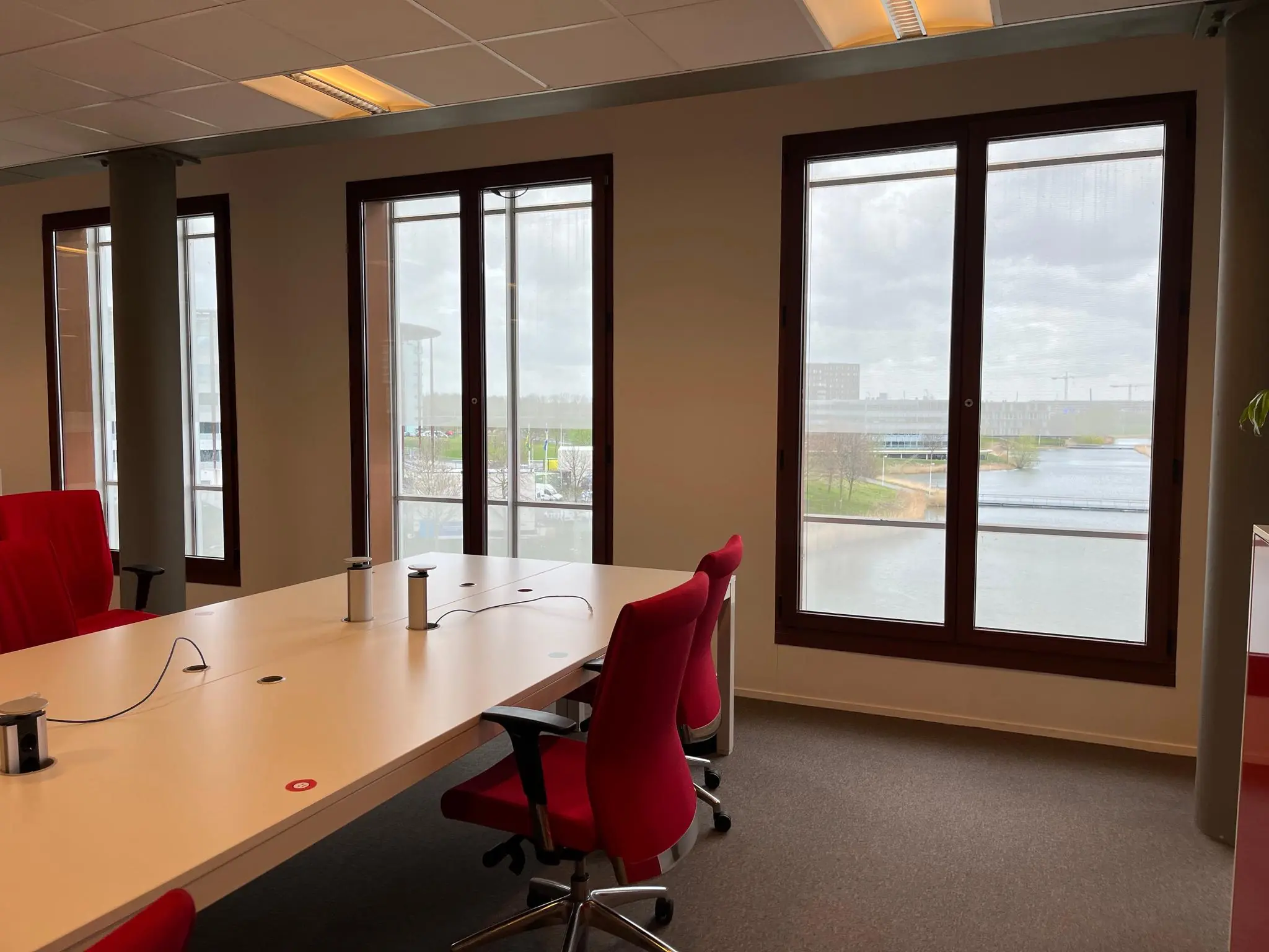 Vergaderruimte aan de Hondiuslaan met rode stoelen, grote ramen en uitzicht op water en moderne gebouwen.