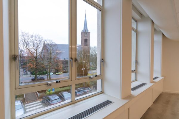 Uitzicht vanuit een modern kantoorraam op de Wassenaarseweg met zicht op een kerk en geparkeerde auto's.