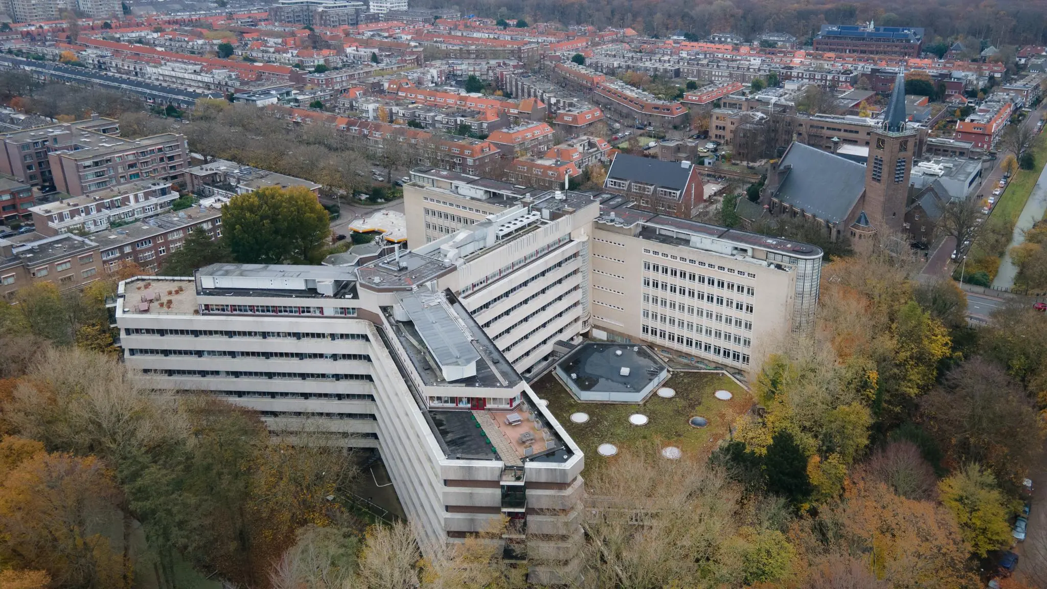 Luchtfoto van het grote kantoorgebouw aan de Wassenaarseweg in Den Haag, omringd door herfstkleurige bomen en woonwijken.