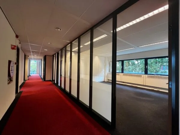 Gang met rood tapijt en glazen wanden die uitkijken op een leeg kantoor aan de Vreeswijksestraatweg.
