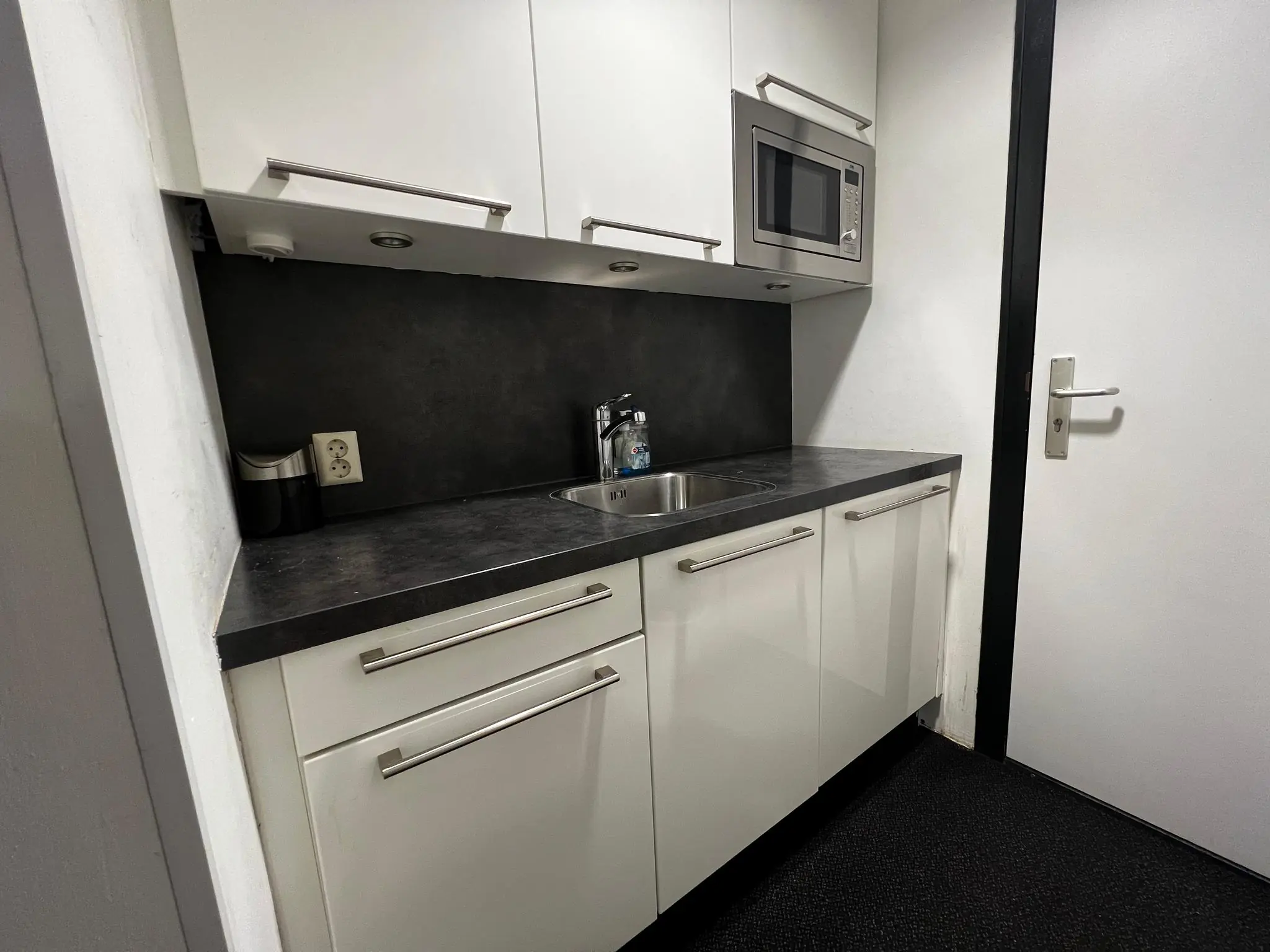 Kleine moderne kitchenette met witte kastjes, zwarte aanrechtblad, ingebouwde magnetron en een spoelbak.