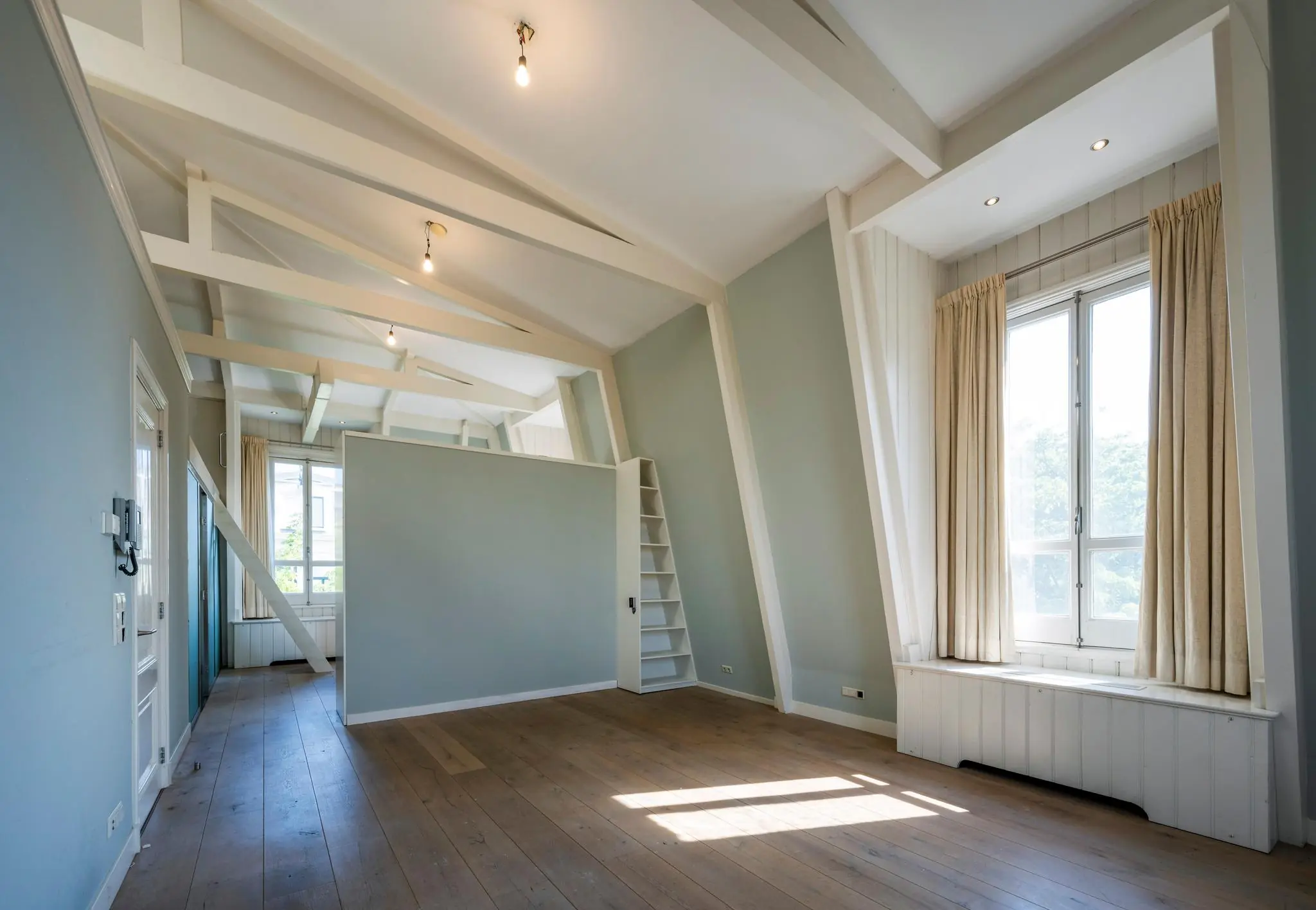Lichte zolderkamer aan de Weteringschans met houten vloer, zichtbare balken en grote ramen met beige gordijnen.