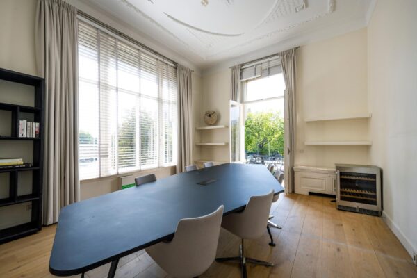 Vergaderruimte aan de Weteringschans met een grote tafel, zes stoelen, hoge ramen en uitzicht op groen.