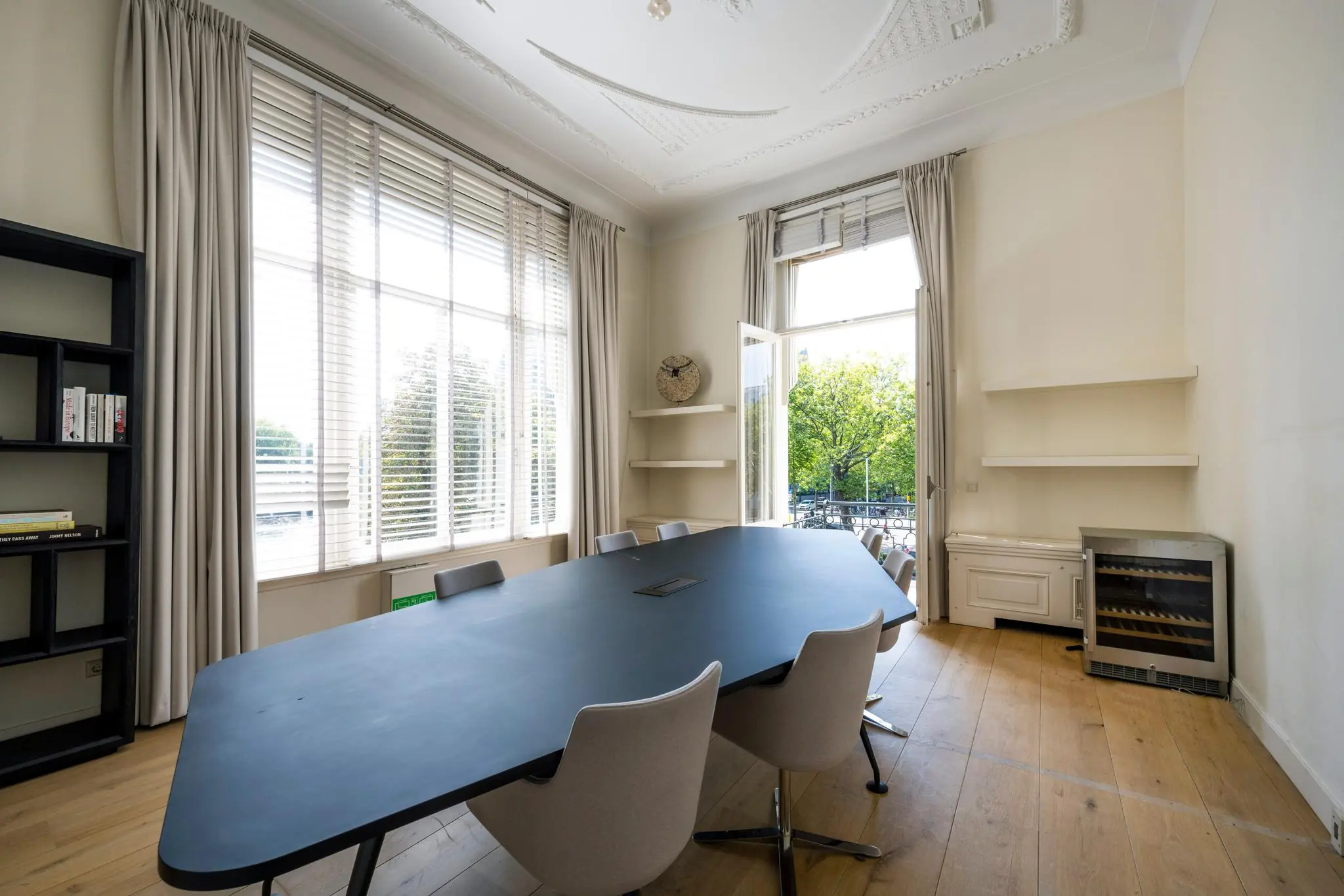 Vergaderruimte aan de Weteringschans met een grote tafel, zes stoelen, hoge ramen en uitzicht op groen.
