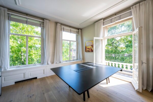 Licht kantoor aan de Weteringschans met houten vloer, groot zwart bureau en openslaande deuren naar een balkon met uitzicht op groene bomen.