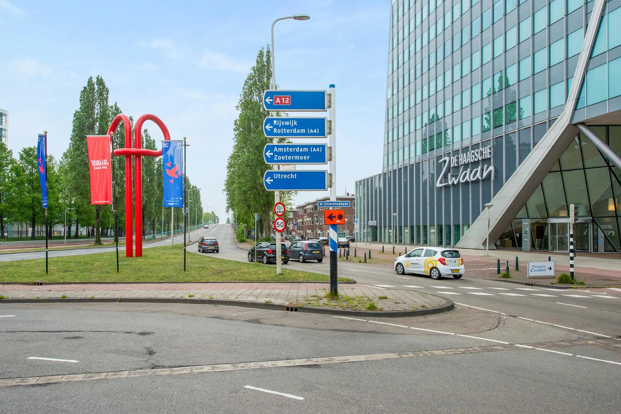 Verkeersplein bij de Schenkkade in Den Haag met het moderne kantoorgebouw De Haagsche Zwaan, opvallende rode kunst en richtingaanwijzers naar A12 en omliggende steden.