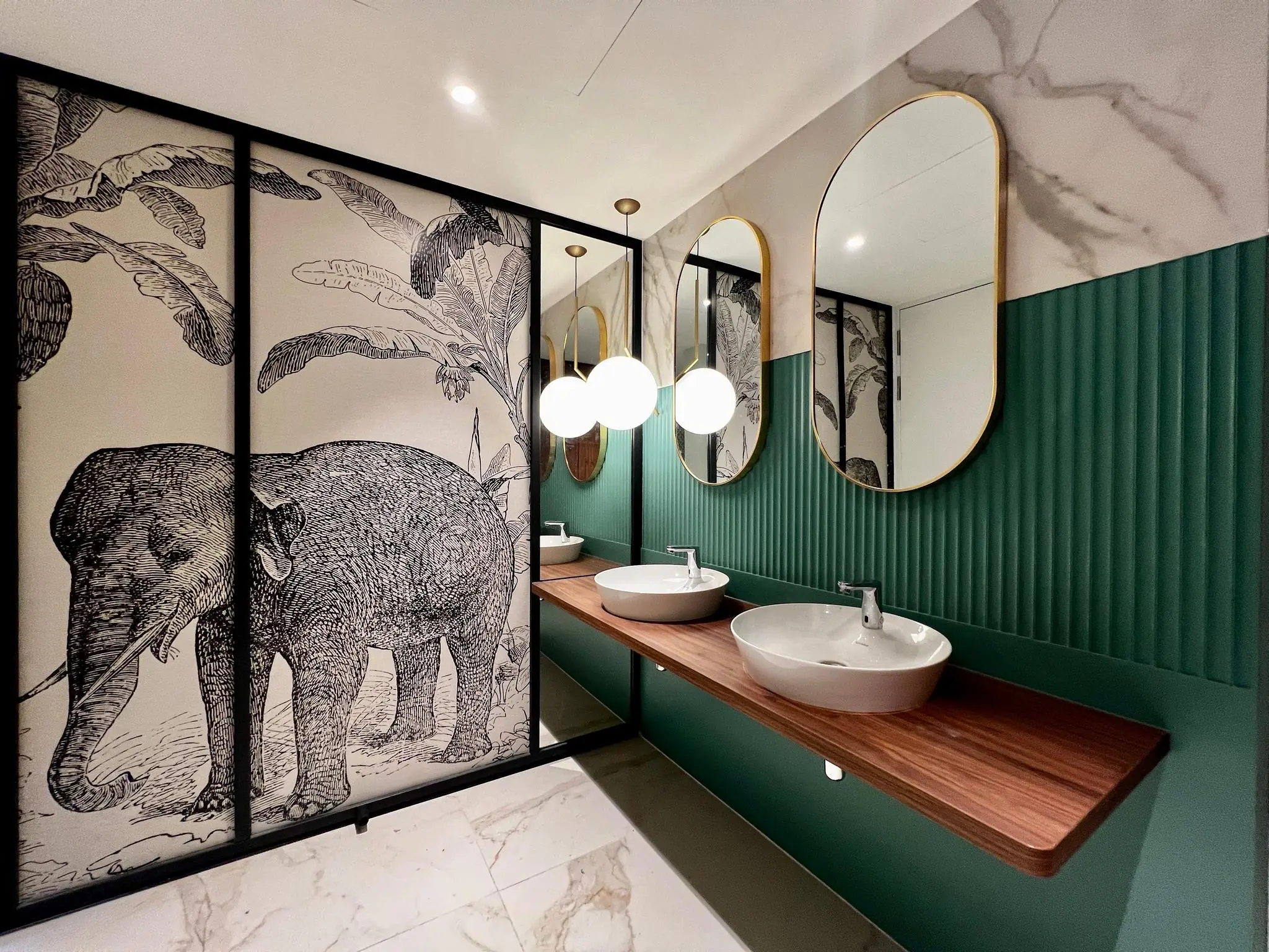 Modern ingerichte toiletruimte met een houten wastafelblad, ronde spiegels, groene wandpanelen en een opvallende wanddecoratie met een getekende olifant.