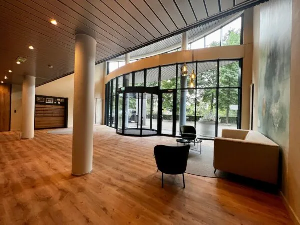 Moderne en ruime entreehal met zitgedeelte, houten vloer en een glazen draaideur die uitkijkt op groen buitengebied.