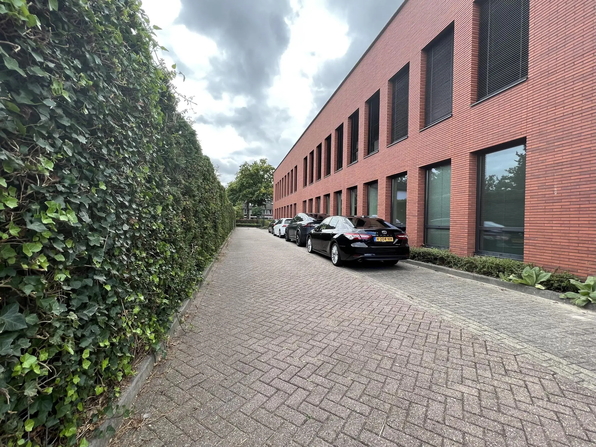 Smalle straat langs een rood bakstenen kantoorgebouw aan de Groenekanseweg, met geparkeerde auto’s en een hoge heg aan de overzijde.