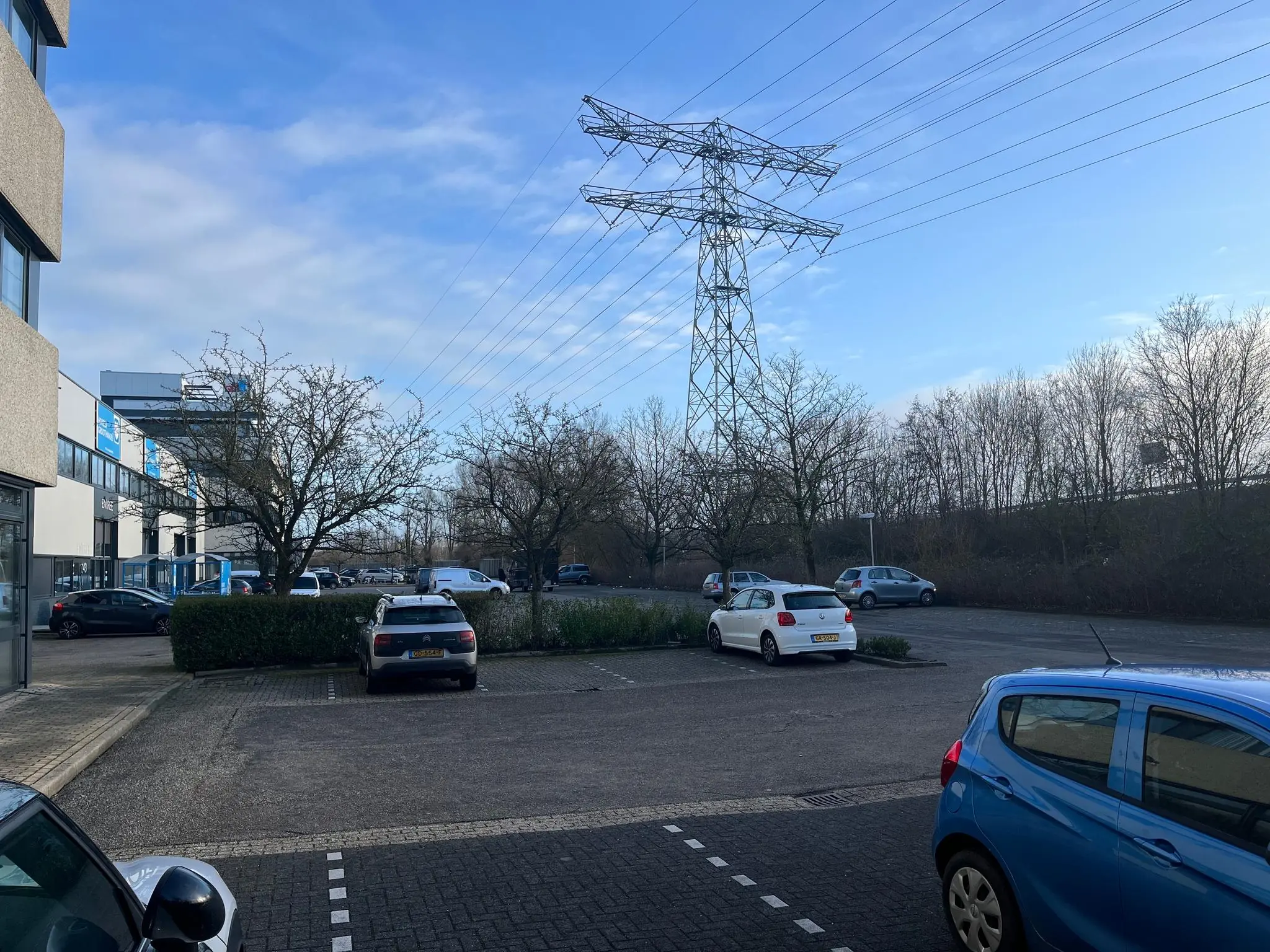 Parkeerterrein aan de Zonnebaan met geparkeerde auto's, bomen en een hoogspanningsmast onder een blauwe lucht.
