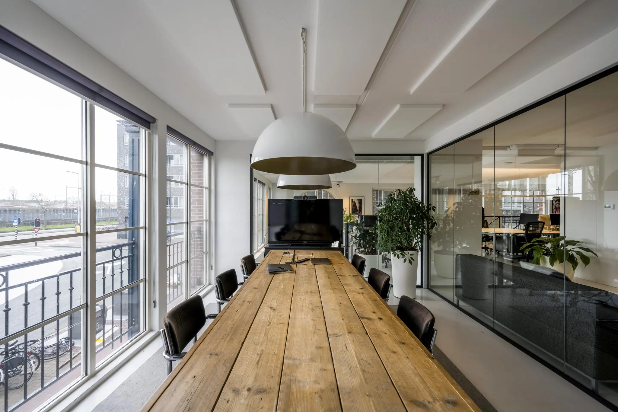 Vergaderruimte aan de Oostelijke Handelskade met een lange houten tafel, grote hanglampen en een tv-scherm, omringd door glazen wanden en uitzicht op de straat.