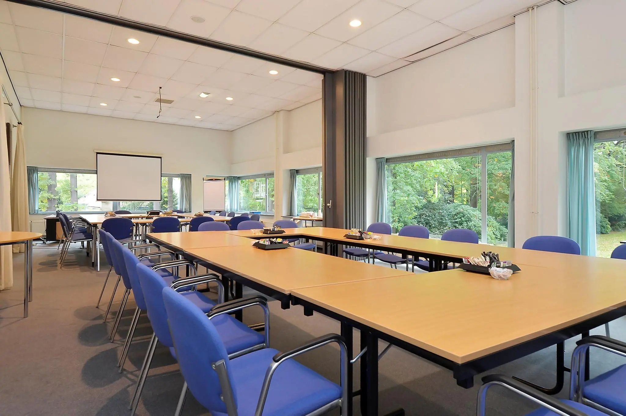 Vergaderzaal aan de Endegeesterstraatweg met opgestelde tafels, blauwe stoelen en uitzicht op groen via grote ramen.