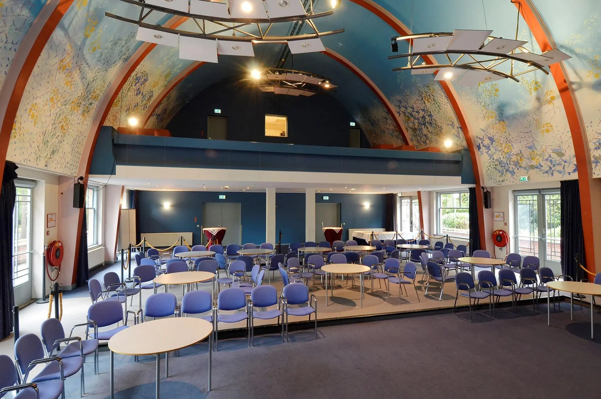 Vergaderzaal met kleurrijk beschilderd gewelfplafond aan de Endegeesterstraatweg, ingericht met ronde tafels en blauwe stoelen.