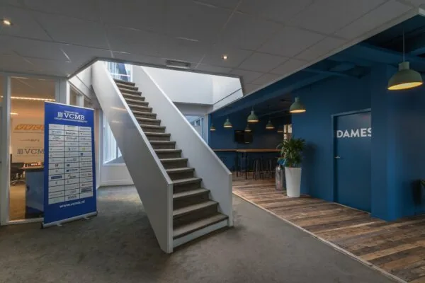 Modern kantoorinterieur met een trap, vergaderruimte van VCMB en een blauwe muur met toiletaanduiding "DAMES".