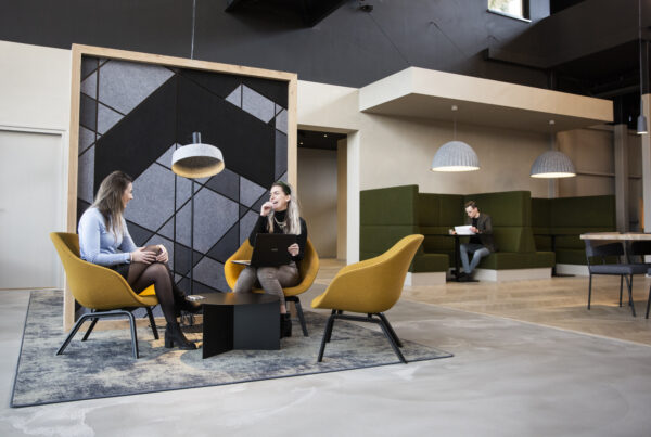 Twee vrouwen voeren een gesprek in een moderne kantoorruimte met gele stoelen en een grafisch wandpaneel op Atoomweg.
