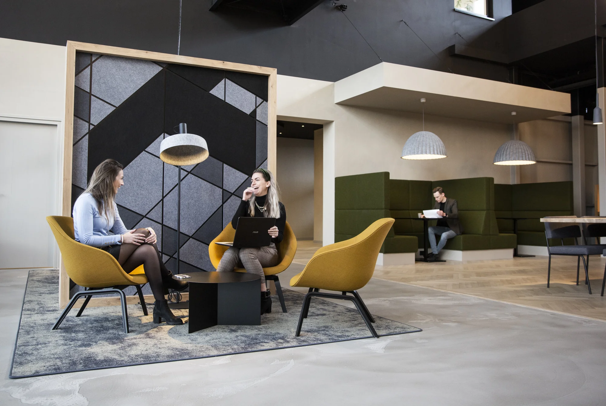 Twee vrouwen voeren een gesprek in een moderne kantoorruimte met gele stoelen en een grafisch wandpaneel op Atoomweg.