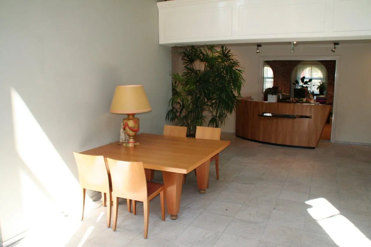 Houten eettafel met vier stoelen en sierlamp in een lichte ruimte met planten en een houten balie op Museumlaan.