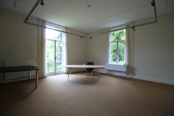Licht kantoor met beige vloerbedekking, een groot bureau bij het raam en uitzicht op groen.