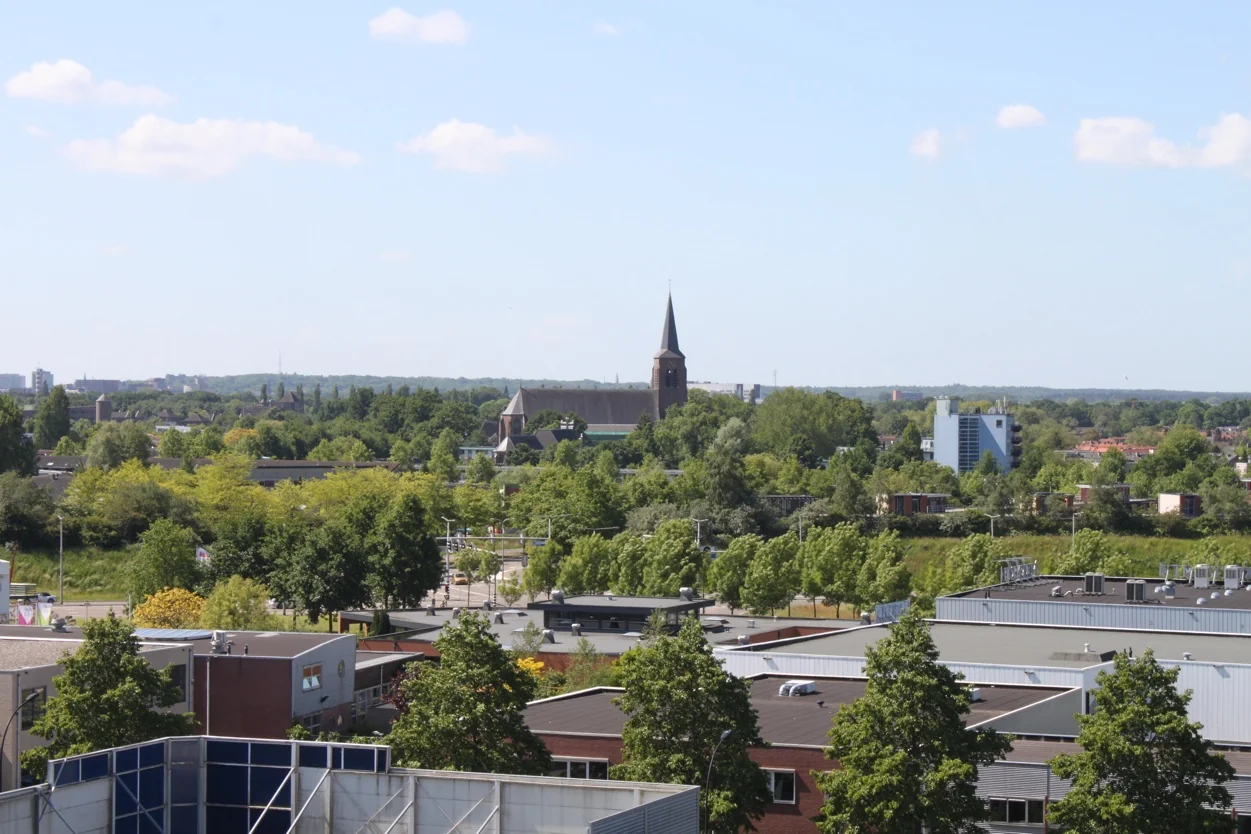 Stadsgezicht met op de achtergrond een kerk met hoge toren, omringd door veel groen en moderne gebouwen.