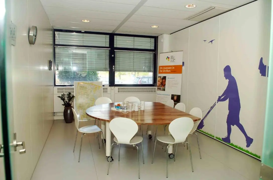 Vergaderruimte met ovale houten tafel, witte stoelen, een muurschildering van een jongen met grasmaaier en een informatief banner over De DrieHen.
