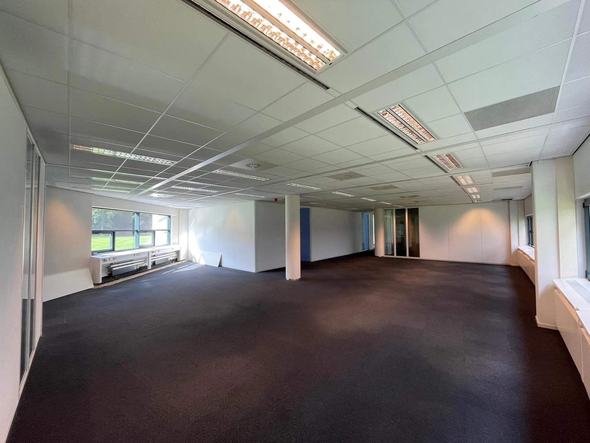 Leeg kantoorpand met systeemplafond, donker tapijt en grote ramen aan beide zijden in gebouw Sparrenheuvel.